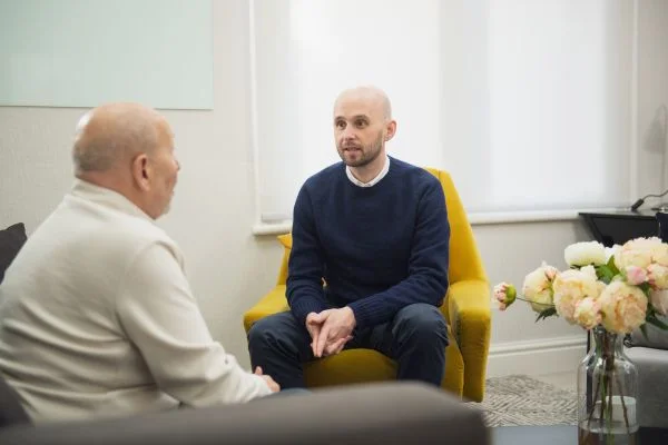 Two men sit facing each other in a bright, softly lit room during what appears to be a counseling or coaching session. In the foreground, an older man with a shaved head, wearing a cream-colored sweater, is seen from behind and side profile, listening attentively. Facing him, a younger bald man with a short beard, dressed in a navy blue sweater over a white collared shirt, leans forward slightly with his hands clasped, speaking earnestly. Between them sits a mustard-yellow armchair. To the right, a glass vase holds a bouquet of pale pink and cream peonies, adding a soft, warm touch to the neutral-toned room with its light walls and sheer curtains.