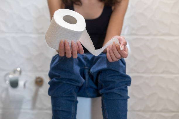 Woman sitting on a toilet holding a roll of toilet paper, jeans pulled down to her knees